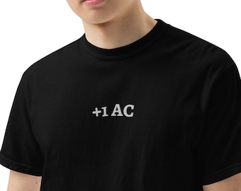 Puede incluir: Camiseta negra con el texto blanco "+1 AC" en el pecho. La camiseta está hecha de un material suave y tiene mangas cortas. El estilo es casual y minimalista.