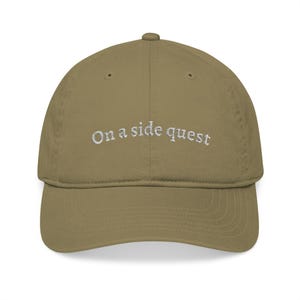 Könnte beinhalten: Khaki-farbene Baseballkappe mit dem gestickten Text "On a side quest" in Weiß. Die Kappe hat einen gebogenen Schirm und ein klassisches Sechs-Panel-Design, geeignet für Freizeitkleidung.