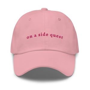 May include: A light pink baseball cap featuring the embroidered text "on a side quest" in a dark pink font across the front panels. The cap has a curved brim, a button on the top, and stitched eyelets for ventilation on each of the six panels.