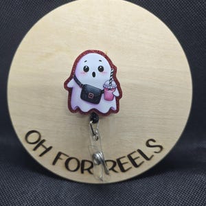 May include: A badge reel featuring a cartoon ghost with a pink drink and a black purse. The ghost has a red glitter outline. The badge reel is attached to a wooden circle with the words "OH FOR REELS" engraved.