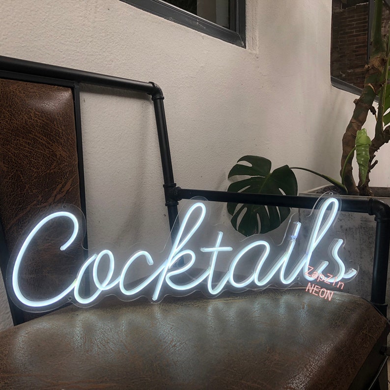 Puede incluir: Letrero de ne&oacute;n blanco con la palabra "Cocktails" en letra cursiva. El letrero est&aacute; montado en un marco de metal negro y est&aacute; iluminado. El letrero est&aacute; en una silla de cuero marr&oacute;n.