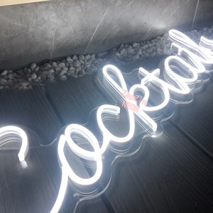 Puede incluir: Un letrero de ne&oacute;n blanco que dice "Cocktail" con un respaldo de acr&iacute;lico transparente. El letrero est&aacute; sobre una superficie de madera.