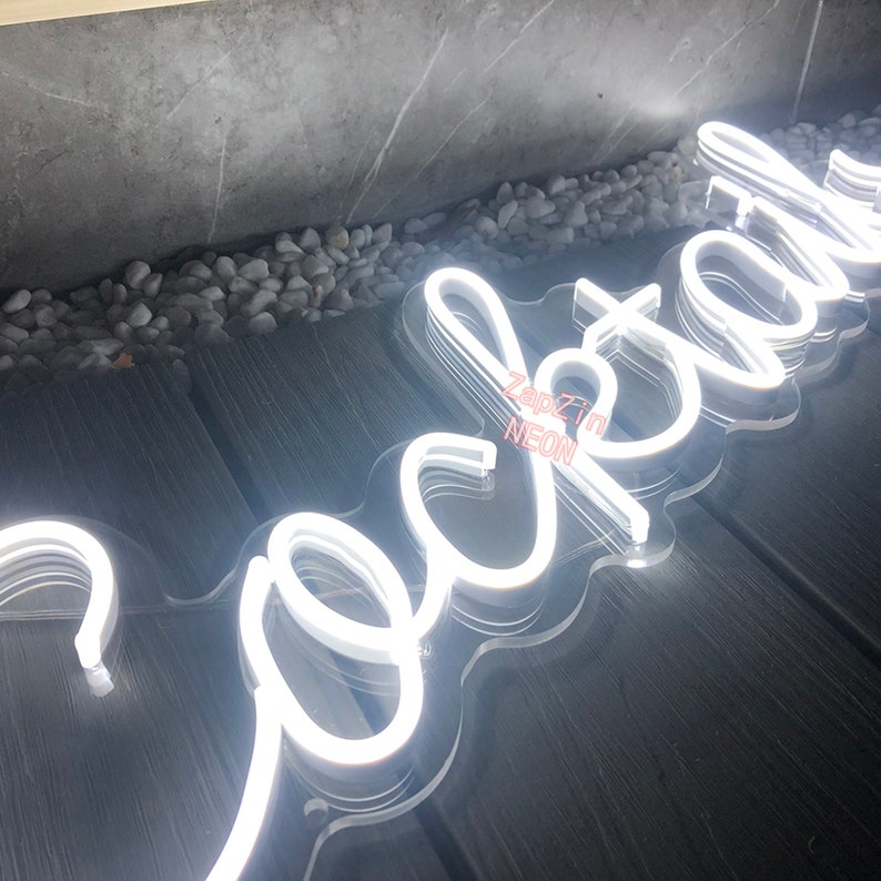 Puede incluir: Un letrero de ne&oacute;n blanco que dice "Cocktail" con un respaldo de acr&iacute;lico transparente. El letrero est&aacute; sobre una superficie de madera.