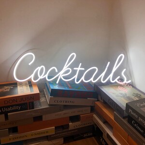 Puede incluir: Letrero de ne&oacute;n blanco con la palabra "Cocktails" en letra cursiva. El letrero est&aacute; sobre una pila de libros.