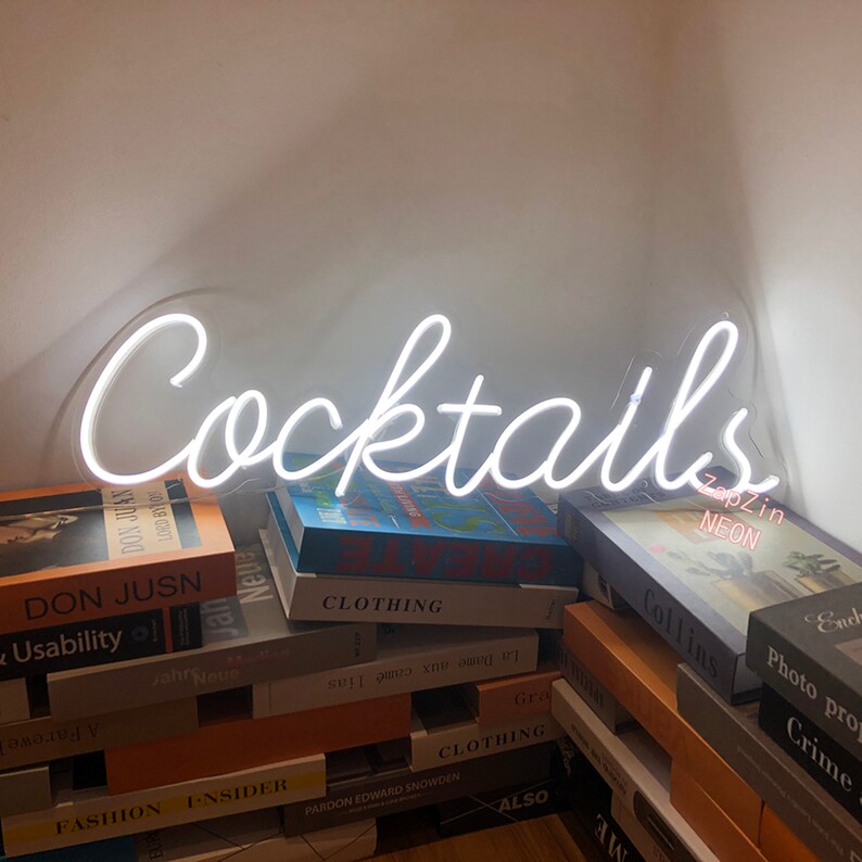 Puede incluir: Letrero de ne&oacute;n blanco con la palabra "Cocktails" en letra cursiva. El letrero est&aacute; sobre una pila de libros.
