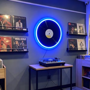 Könnte beinhalten: Innenraum eines Plattenladens mit einer blauen Neon-Vinylplatte an der Wand. Schwarze Regale zeigen Albumcover. Ein Plattenspieler steht auf einem Holztisch mit schwarzem Metallrahmen. Weitere Alben sind sichtbar.
