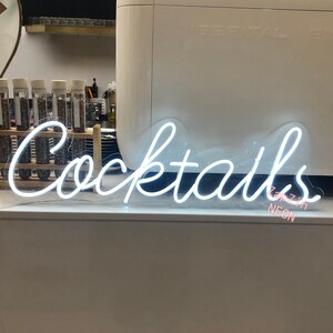 Puede incluir: Letrero de ne&oacute;n blanco con la palabra "Cocktails" en una fuente cursiva. El letrero est&aacute; iluminado y tiene un respaldo de pl&aacute;stico transparente. El texto "Zappzin Neon" es visible en rosa debajo del letrero.