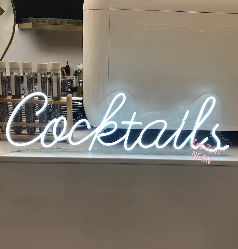 Puede incluir: Letrero de ne&oacute;n blanco con la palabra "Cocktails" en una fuente cursiva. El letrero est&aacute; iluminado y tiene un respaldo de pl&aacute;stico transparente. El texto "Zappzin Neon" es visible en rosa debajo del letrero.