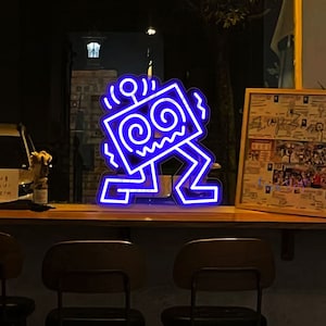 May include: A neon blue light fixture depicting a stylized figure with a square head, spiral eyes, and zig-zag arms and legs. The figure is lit against a dark backdrop, possibly in a bar or restaurant.