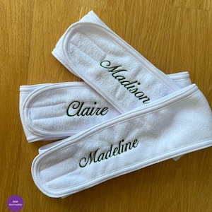 May include: Three white terry cloth spa headbands with embroidered names in green script: "Madison", "Claire", and "Madeline". The headbands are folded and arranged on a wooden surface.