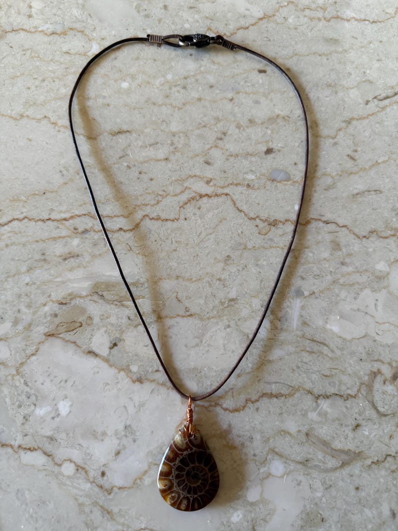 Puede incluir: Collar con colgante en forma de l&aacute;grima con un amonita fosilizada pulida. El colgante es marr&oacute;n y crema, con un patr&oacute;n en espiral. El collar tiene un cord&oacute;n marr&oacute;n oscuro y alambre de cobre.