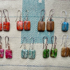 Puede incluir: Una colección de pendientes hechos a mano con cuentas de piedra rectangulares en varios colores, incluyendo rojo, marrón, azul, rosa, verde y beige. Cada par tiene un patrón único y está alambrado con alambre plateado o cobrizo.