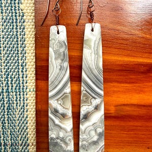 May include: A pair of long, rectangular earrings with a marbled pattern in shades of gray, white, and tan. The earrings are suspended from copper-colored ear wires. The earrings are displayed on a wooden surface.