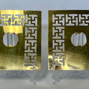 Op de afbeelding: Twee vierkante, goudkleurige metalen decoratieve panelen met een geometrisch uitgesneden ontwerp. Elk paneel heeft een cirkelvormige opening en wordt gepresenteerd op een transparante plastic standaard. De panelen hebben een vintage esthetiek.
