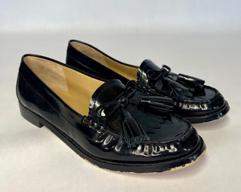 May include: Pair of glossy black leather loafers with tassel details. The shoes have a classic design with a rounded toe and a low heel. The loafers are displayed on a white background.