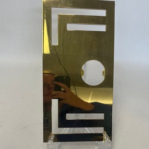Op de afbeelding: Een rechthoekige, goudkleurige metalen plaat met het woord "FEEL" uitgesneden. De plaat wordt ondersteund door een transparante plastic standaard. Het oppervlak reflecteert de omgeving.