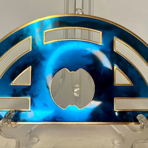 Op de afbeelding: Een halfrond decoratief stuk met een blauw en goud ontwerp. Het stuk heeft een metallic blauwe afwerking met gouden randen en geometrische uitsparingen. Het midden heeft een spiegelend element. Het stuk wordt op een transparante standaard gepresenteerd.