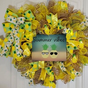 May include: A yellow and green summer wreath with a sign that says "Summer vibes". The wreath is decorated with pineapples, ribbons, and mesh.