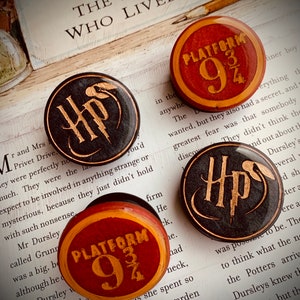 May include: Three round Harry Potter themed pins. One pin features the Hogwarts crest in gold on a black background. Another pin features the Platform 9 3/4 sign in gold on a red and yellow background. The third pin is the same as the second pin but with a black and gold background.