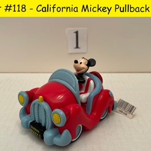 May include: Red pullback toy car with a blue roof and Mickey Mouse driving. The car has a yellow grill and headlights. The car has the words "California Mickey 1" on the side. The car is on a white surface.