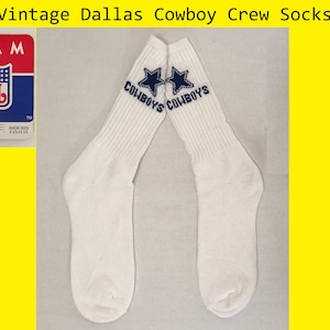 May include: A pair of white crew socks with the Dallas Cowboys logo and the words "COWBOYS" on each sock. The socks have a blue star with a white outline on each sock. The socks are new and unused.