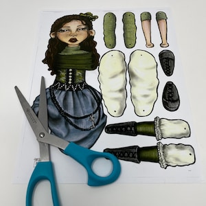 May include: A paper doll craft kit with a hand-drawn illustration of a woman in a green and blue dress. The kit includes cut-out pieces for the doll's body, clothing, and accessories. A pair of blue-handled scissors is placed on top of the paper.