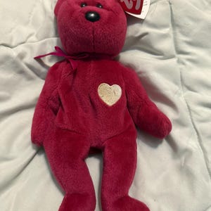 Puede incluir: Un oso de peluche magenta con un parche dorado en forma de corazón en el pecho. El oso tiene una nariz negra, una cinta magenta y una etiqueta Ty. El peluche mide aproximadamente 20 cm de alto. Un juguete clásico para niños.