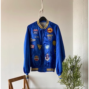 Op de afbeelding: Een blauwe satijnen bomberjack met gele en witte geribbelde boorden aan de kraag, manchetten en zoom. De jas is versierd met verschillende geborduurde patches, waaronder een Route 66-bord, een smiley, een anker en een Benelli-logo.