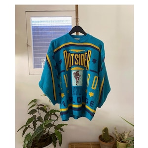 May include: A blue, yellow and black knitted jumper with the words "Outsider Worldwide" and a graphic of a person in a basketball pose. The jumper has a star pattern.