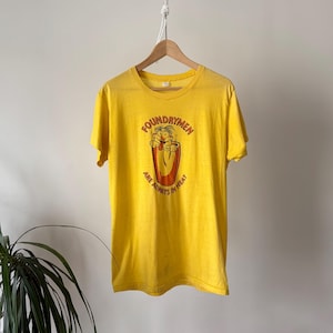 Puede incluir: Camiseta amarilla con un gráfico de un personaje de dibujos animados dentro de un recipiente, con el texto "FOUNDRYMEN ARE ALWAYS IN HEAT". La camiseta está colgada de una percha de madera.