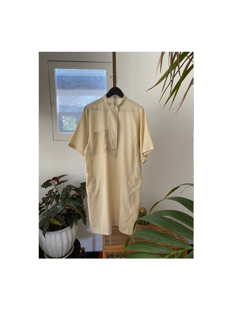 May include: A pale yellow short-sleeved shirt with a mandarin collar and a single patch pocket. The shirt is hanging on a black hanger.