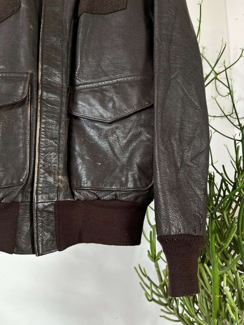 Op de afbeelding: Een bruine leren bomberjack met een geribbelde gebreide kraag, manchetten en tailleband. De jas heeft een ritssluiting en twee klepzakken.