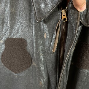 Op de afbeelding: Een bruin leren jack met een ritssluiting en een bruin fleece-patch aan de linkerkant van de borst.