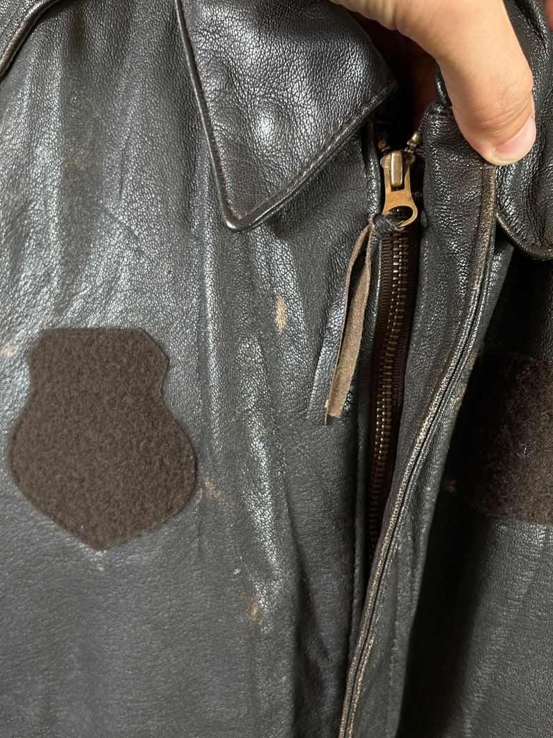 Op de afbeelding: Een bruin leren jack met een ritssluiting en een bruin fleece-patch aan de linkerkant van de borst.