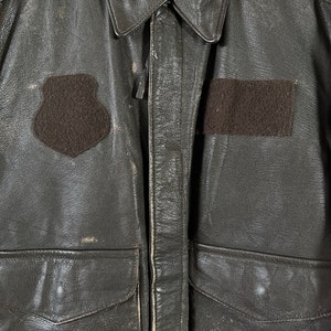 Op de afbeelding: Een bruin leren jack met een ritssluiting en twee klepzakken. Het jack heeft een patch op de linkerborst met de tekst "VIREX LTD.  MIL-J-558-620 AF FLIGHT JACKET LOT 102".