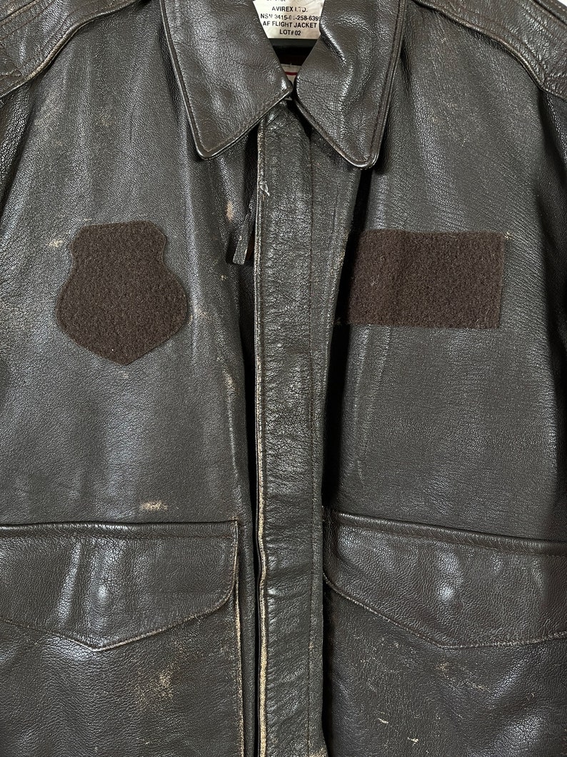 Op de afbeelding: Een bruin leren jack met een ritssluiting en twee klepzakken. Het jack heeft een patch op de linkerborst met de tekst "VIREX LTD.  MIL-J-558-620 AF FLIGHT JACKET LOT 102".