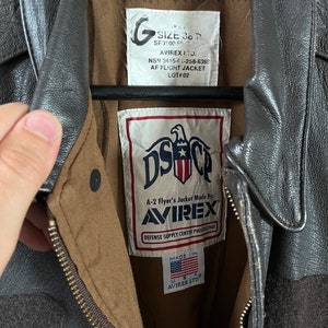 Op de afbeelding: Een bruin leren A-2 vluchtjack met een ritssluiting. Het jack heeft een label met de tekst "A-2 Flyer's Jacket Made By: AVIREX" en "Defense Supply Center Philadelphia". Het label vermeldt ook "Made in USA" en "AVIREX LTD."