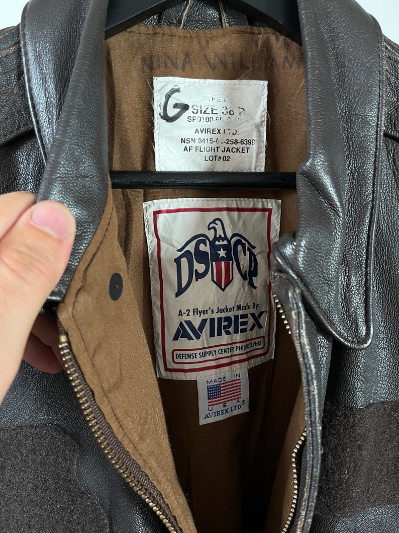 Op de afbeelding: Een bruin leren A-2 vluchtjack met een ritssluiting. Het jack heeft een label met de tekst "A-2 Flyer's Jacket Made By: AVIREX" en "Defense Supply Center Philadelphia". Het label vermeldt ook "Made in USA" en "AVIREX LTD."