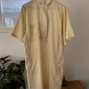 May include: A pale yellow short-sleeved collared dress with a button-up front and a single pocket on the left side. The dress has a relaxed fit and a slightly longer length. The label on the dress reads "Roger Gallet".