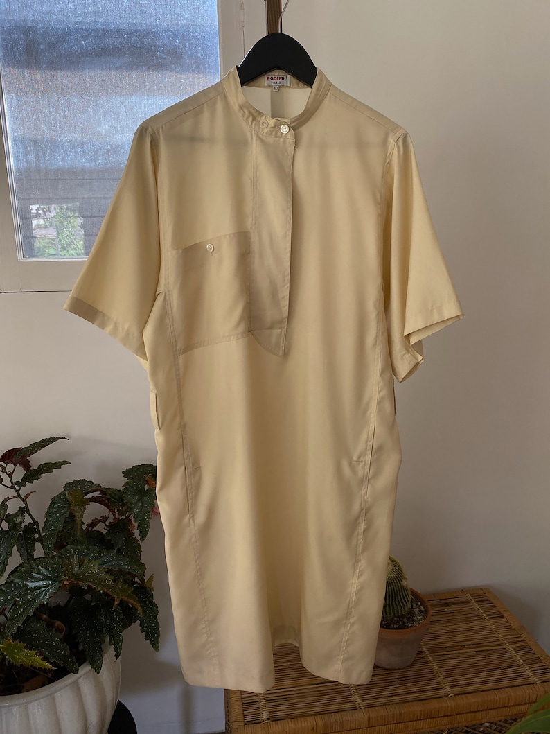 May include: A pale yellow short-sleeved collared dress with a button-up front and a single pocket on the left side. The dress has a relaxed fit and a slightly longer length. The label on the dress reads "Roger Gallet".