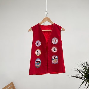 Op de afbeelding: Rode, mouwloze vest met verschillende geborduurde patches. De patches bevatten afbeeldingen van mensen, gebouwen en andere ontwerpen.