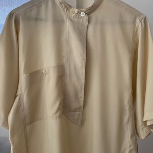 May include: A light beige short-sleeved shirt with a mandarin collar and a single button closure. The shirt has a patch pocket on the left side and a long, narrow flap that extends from the collar to the hem. The shirt has a "Rodier Paris" label with the size "40" printed on it.