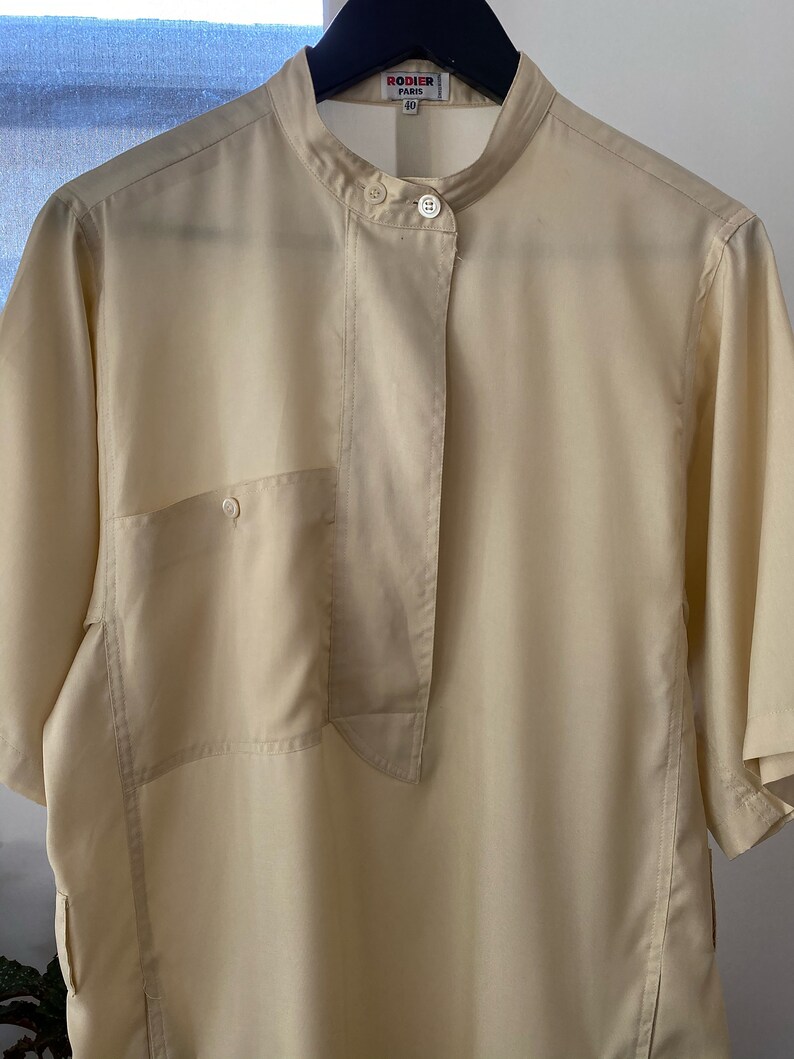 May include: A light beige short-sleeved shirt with a mandarin collar and a single button closure. The shirt has a patch pocket on the left side and a long, narrow flap that extends from the collar to the hem. The shirt has a "Rodier Paris" label with the size "40" printed on it.