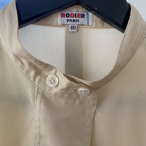 May include: A beige button-up shirt with a mandarin collar and two white buttons. The shirt has a label that reads "Rodier Paris" and "40".
