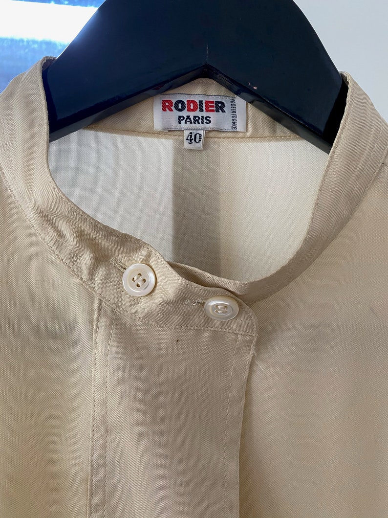 May include: A beige button-up shirt with a mandarin collar and two white buttons. The shirt has a label that reads "Rodier Paris" and "40".