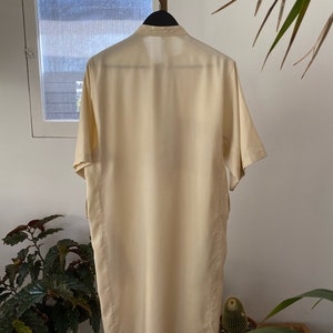 May include: A long, cream-colored shirt with a collar and short sleeves. The shirt is hanging on a hanger and is made of a smooth, silky fabric.