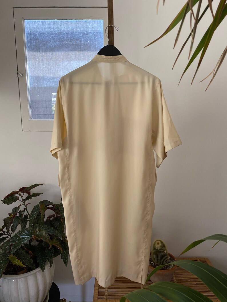May include: A long, cream-colored shirt with a collar and short sleeves. The shirt is hanging on a hanger and is made of a smooth, silky fabric.
