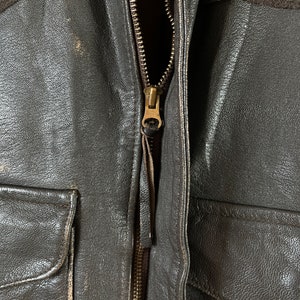 Op de afbeelding: Close-up van een bruin leren jack met een messing rits. De rits is gedeeltelijk open, waardoor de bruine voering van het jack zichtbaar is.