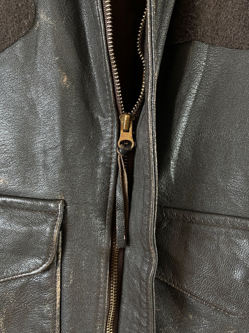 Op de afbeelding: Close-up van een bruin leren jack met een messing rits. De rits is gedeeltelijk open, waardoor de bruine voering van het jack zichtbaar is.