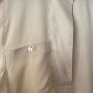 May include: A close-up of a beige button-down shirt with a single patch pocket on the left side. The shirt has a mandarin collar and a single button closure at the neck.